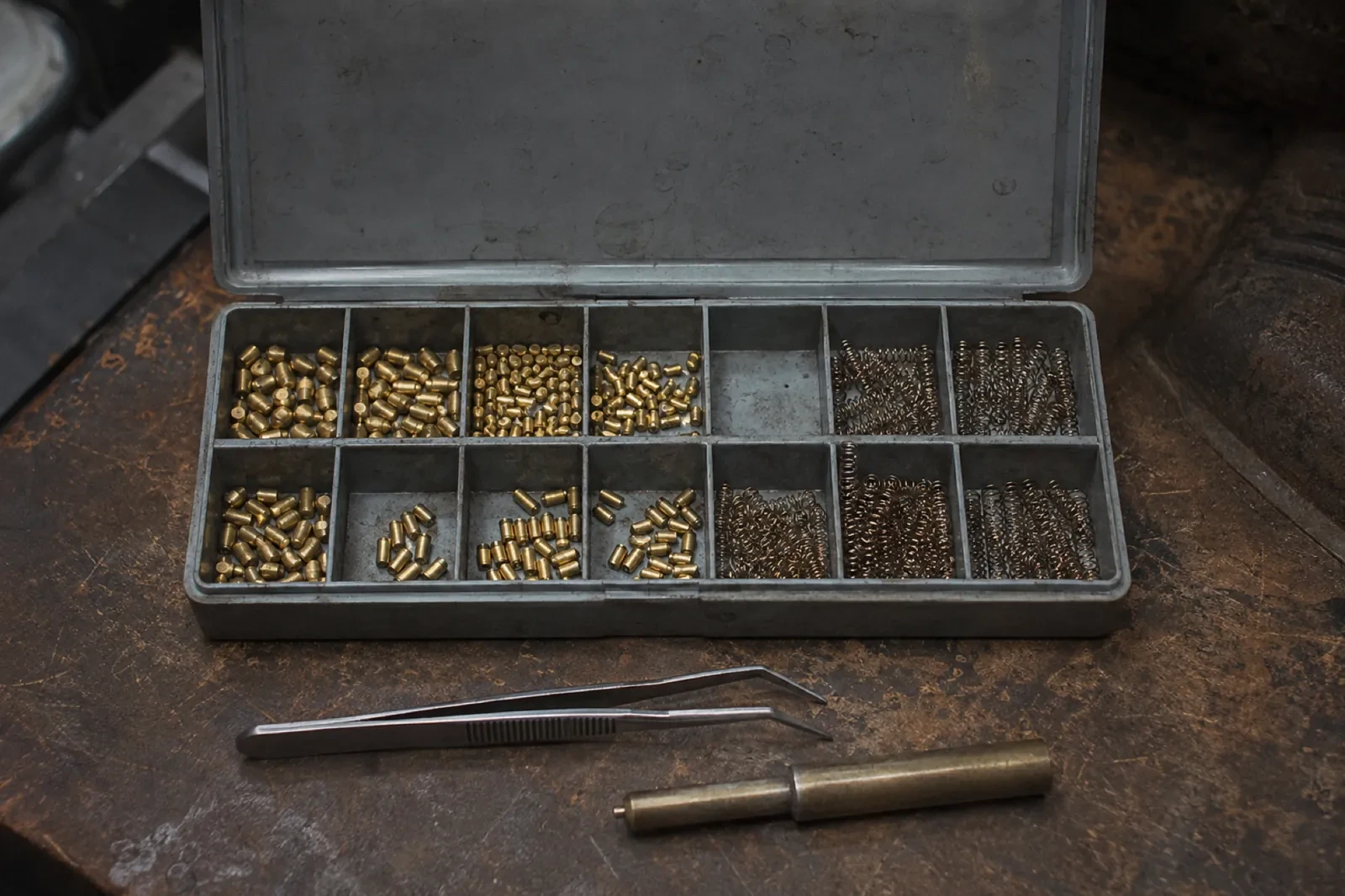 Open metal case with brass pins and coiled springs sorted in small sections, with tweezers and a brass tool in front. — Port Union